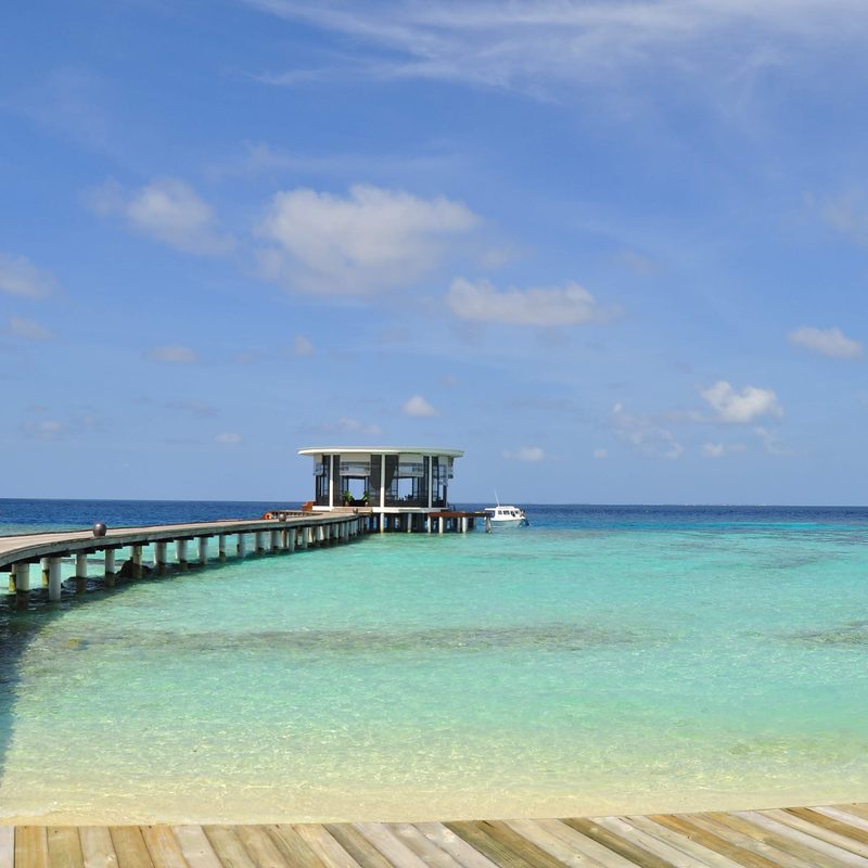 A timber jetty running out to an overwater pavilion on a turquoise lagoon.