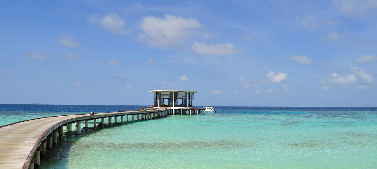 A timber jetty running out to an overwater pavilion on a turquoise Maldivian lagoon.