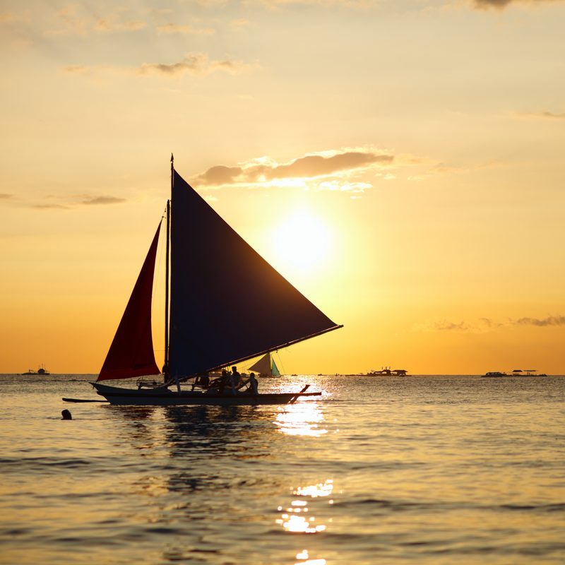 A paraw sailboat silhouetted against a gold sunset over calm sea.